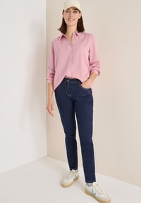 Helle rosa Button-Up-Bluse mit hochgerollten Ärmeln, kombiniert mit dunkelblauen Skinny Jeans und cremefarbenen Sneakers mit blauen Akzenten.