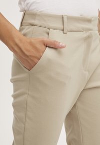 Beige housut, joissa on sileä pinta, sivutasku ja siistit etupleatit. Vyötärö on litteä ilman näkyvää metalliosaa.