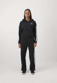 Svart zip-up huvtröja och byxset med tre vita ränder på ärmar och sidor, med en broderad logotyp på bröstet och elastiska muddar.