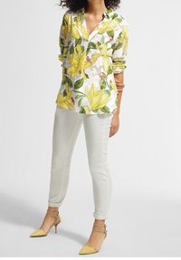 Blouse blanche à manches longues avec un motif floral jaune, associée à un pantalon crème ajusté et des talons pointus jaunes avec brides à la cheville.