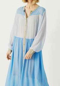 Abito leggero a maniche lunghe in tessuto trasparente bianco e blu, con una chiusura frontale con bottoni e dettagli beige, e una gonna fluente.