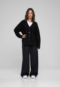 Zwarte oversized cardigan met een kabelpatroon, V-hals en sluiting met zes knopen, gecombineerd met zwarte broek met wijde pijpen en witte schoenen.