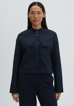 Vrouw met lang zwart haar draagt een donkerblauwe jas met knopen en twee grote zakken aan de voorkant, gecombineerd met een bijpassende broek, en staat voor een effen achtergrond.