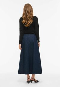 Kobieta z długimi falującymi włosami, ubrana w czarną bluzkę z długim rękawem, granatową spódnicę midi w kształcie litery A oraz czarne buty na niskim obcasie, zwrócona tyłem.