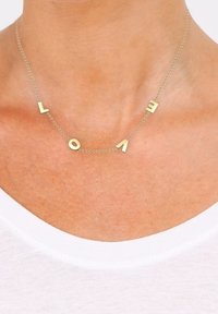 Collier délicat en chaîne d'or avec des breloques en forme de lettres dorées éparpillées formant le mot "LOVE", porté sur un cou nu au-dessus d'une chemise blanche.