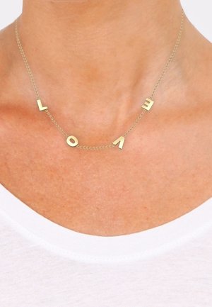 Collier délicat en chaîne d'or avec des breloques en forme de lettres dorées éparpillées formant le mot "LOVE", porté sur un cou nu au-dessus d'une chemise blanche.
