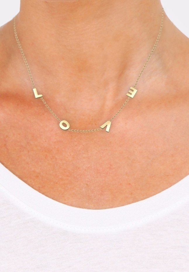 Collier délicat en chaîne d'or avec des breloques en forme de lettres dorées éparpillées formant le mot "LOVE", porté sur un cou nu au-dessus d'une chemise blanche.