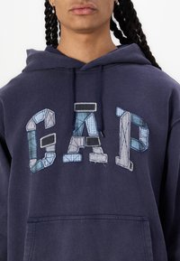 Zilais kapuci džemperis ar raiboni "GAP" logo, kas veidots no dažādām zilajām raibajām un denima tekstūrām, izpildīts ar kontrastējošām šuvēm un priekšējo kabatu.