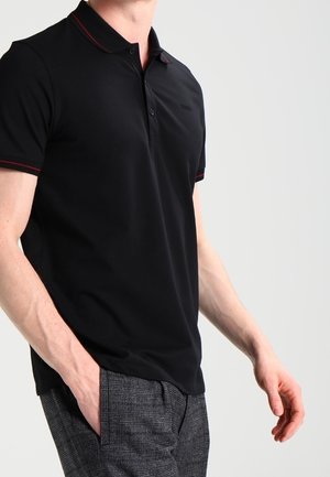 Mand iført sort, kortærmet poloshirt med rød kant og grå ternede bukser, stående med den ene hånd i lommen mod en ensfarvet baggrund.