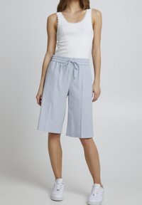 Žena v bílém bezrukávovém topu, volných světle modrých šortkách ke kolenům na šňůrku a bílých teniskách na jednobarevném šedém pozadí.