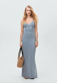 Ljusblå stickad maxiklänning med V-ringning, figurnära byst, virkade detaljer och en slät textur. Modellen håller en liten vävd väska.