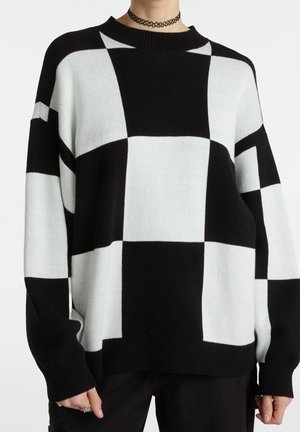 Osoba nosząca czarno-biały, oversize, w kratę sweter z prążkowanymi mankietami i dołem, połączony z czarnymi spodniami oraz czarnym naszyjnikiem typu choker.