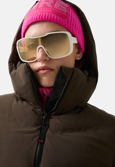 Braune gepolsterte Jacke mit hohem Kragen, ausgestattet mit einem schwarzen Reißverschluss. Kombiniert mit einer lebhaft pinken Strickmütze und übergroßen, weiß getönten Brillen.