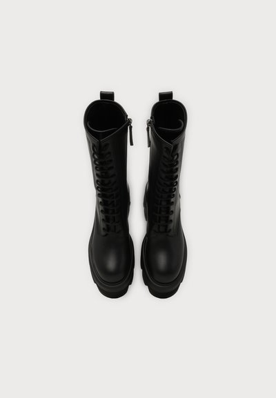 Paire de bottes de combat noires à lacets avec fermetures éclair latérales et semelles épaisses, vue de dessus sur un fond uni clair.
