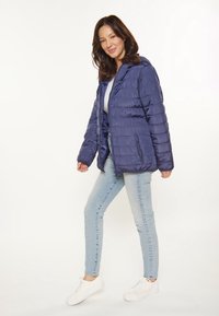 Chaqueta acolchada azul marino con capucha, que presenta un diseño acolchado y bolsillos laterales con cremallera. Combinada con unos vaqueros azul claros y zapatillas blancas.