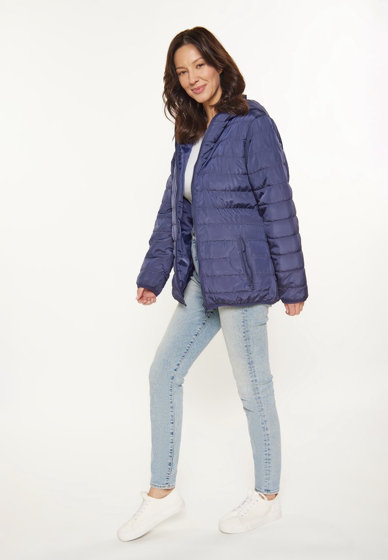 Chaqueta acolchada azul marino con capucha, que presenta un diseño acolchado y bolsillos laterales con cremallera. Combinada con unos vaqueros azul claros y zapatillas blancas.