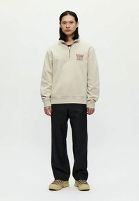 Hombre vistiendo un sudadera de media cremallera color beige con logo, pantalones negros y zapatillas beige, de pie contra un fondo blanco liso.