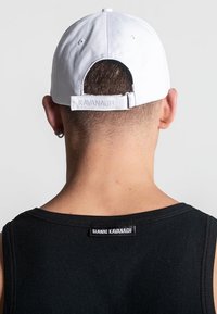 Bijela bejzbol kapa s trakicom straga i utisnutim logom "KAVANAGH". Crni pamučni top bez rukava s vidljivom oznakom brenda na izrezu vratu.