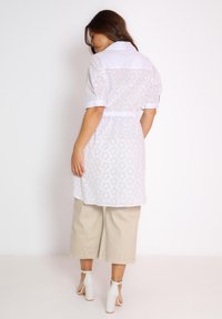 Witte, geborduurde shirtdress met een kraag, korte mouwen en een getailleerde taille. Gecombineerd met beige wijde broek en witte sandalen met hak.