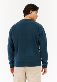 Blauwe fleece sweater met ronde hals, ribgebreide boorden en onderrand. Heeft een gladde textuur en een ontspannen pasvorm. Draagt over lichtgekleurde cargobroek.