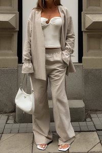 Bež laneni komplet s širokimi hlačami in sproščenim jopičem. Prekrivni bel top ima sprednji zasuk. Dodatki vključujejo belo torbico.