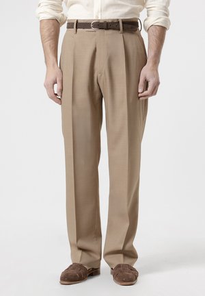 Hombre con pantalones plisados beige, cinturón de cuero marrón, mocasines de ante marrón y una camisa de manga larga color crema con las mangas remangadas.