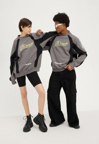 Deux personnes portant des sweatshirts "Silver Racer" gris et noirs se tiennent face à face, l'une avec un short noir et des bottes, l'autre avec un pantalon noir et des lunettes de soleil.