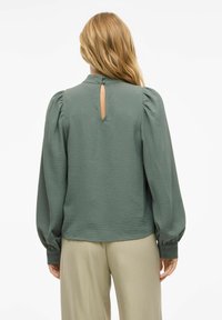 Blouse verte texturée avec des manches bouffantes et une fermeture en clé à l'arrière, assortie à un pantalon beige clair.