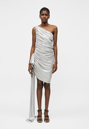 Žena v lesklej striebornej šatke na jedno rameno s nariasením, dlhým pásom a čiernych otvorených podpätkoch stoji na jednoduchej pozadí.