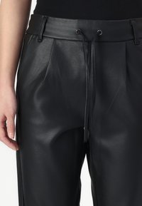 Gros plan d'une personne portant un pantalon noir en similicuir avec des plis, des passants de ceinture et un cordon à la taille, associé à un haut noir.