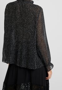 Blouse noire transparente avec des petites taches blanches, encolure à volants, manches longues et ourlet froncé et fluide. Tissu léger et texturé.