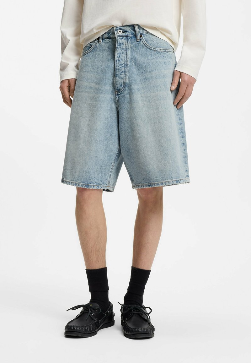 Personne portant un short en jean bleu clair, des chaussettes noires et des chaussures noires à lacets, debout devant un fond blanc uni.