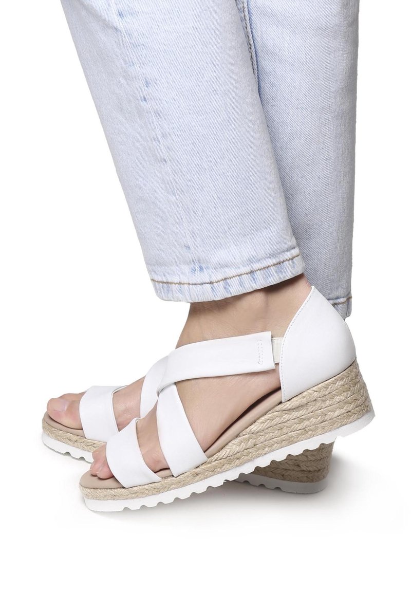 Pieds portant un jean bleu clair et des sandales compensées blanches avec des semelles en corde tressée et des lanières croisées.