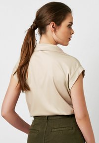 Beige Bluse mit kurzen Ärmeln, Kragen und umgeschlagenen Bündchen, kombiniert mit olivgrünen Hosen mit Gesäßtaschen. Glattes, leichtes Material.