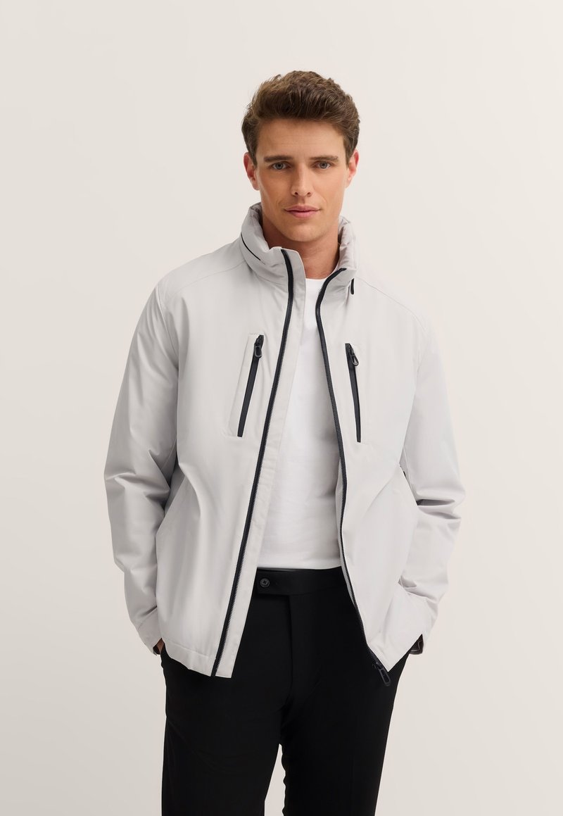 Helle graue Jacke mit hohem Kragen, schwarzen Reißverschlüssen und zwei Vordertaschen. Trägt man über einem weißen Hemd und schwarzen Hosen.