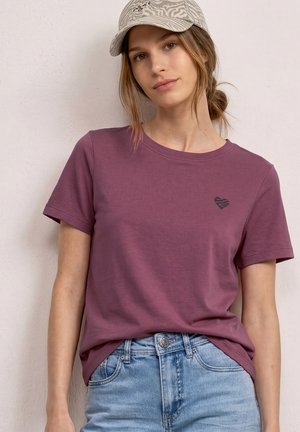 Ung kvinde iført en mullvadsfarvet t-shirt med en lille hjerte-logo og lyse denimjeans, stylet med en beige mønstret baseballkasket.