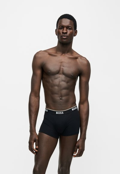 Homme musclé debout, portant un boxer noir avec le logo BOSS sur la ceinture, sur un fond blanc uni.