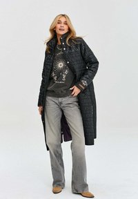 Pikowana czarna długa kurtka z wysokim kołnierzem, w połączeniu z ciemnym sweatshirtem z grafiką, jasnoszarymi dżinsami i beżowymi botkami za kostkę.