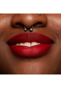 Matt röd läppstift på fylliga läppar, som framhäver den fina texturen och släta finishen. Silver septumpiercing ovanför ett framträdande leende på mörk hud.