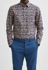 Košile na knoflíky s vícero barevným květinovým vzorem na tmavém pozadí; v kombinaci s úzkými modrými kalhotami, vytvářejícími strukturovaný vzhled.