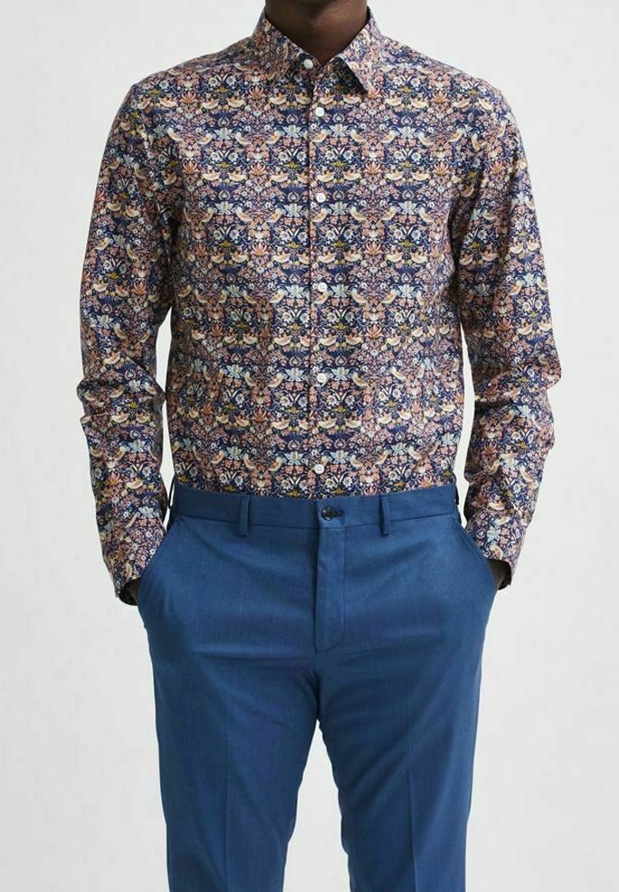 Košile na knoflíky s vícero barevným květinovým vzorem na tmavém pozadí; v kombinaci s úzkými modrými kalhotami, vytvářejícími strukturovaný vzhled.
