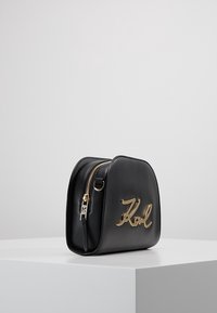 Czarna skórzana torebka o zaokrąglonym kształcie, z złotym metalowym logo "Karl", bocznym zamkiem błyskawicznym i gładką fakturą. Umieszczona na białej powierzchni ekspozycyjnej.