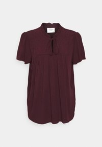 Burgunderfarbene Bluse mit kurzen Ärmeln, rundem Ausschnitt, gefältelter Vorderseite, Bindeschluss und einem strukturierten Punktmuster. Weiche Stoffoberfläche.