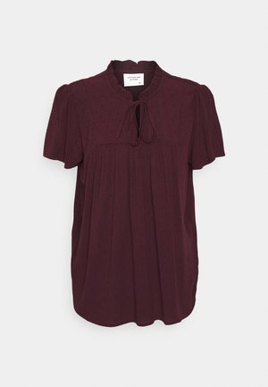Burgundy kortærmet bluse med rund hals, plisseret front, båndlukning og tekstureret prikket mønster overalt. Blødt stoffinish.