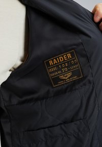 Črn prešiti telovnik z rjavim znakom z besedilom: "RAIDER," "ISSUE: 502/00," in "PME LEGEND." Gladek material in funkcionalni žepi.