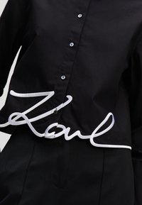 Zwarte button-up shirt met witte geborduurde "Karl" letters aan de zoom, strakke stof en getextureerde accenten.