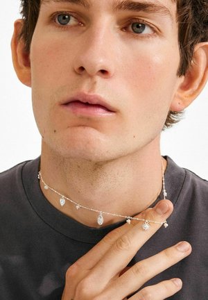 Silver choker with various charms, including leaf and star shapes, featuring a delicate chain and small beads against a gray shirt.
