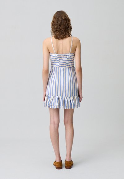 Abito estivo a righe con spalline sottili, caratterizzato da righe verticali blu e crema, una schiena arricciata e un orlo con volant. Indossato con scarpe in suede marroni.