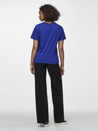 Göndör hajú nő, háttal nézve, kék pólóban, fekete bőszárú farmerral és fekete-fehér tornacipőben, egyszerű háttér előtt.