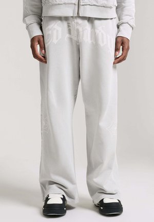 Osoba nosząca jasnoszare, luźne spodnie dresowe z delikatnym gotyckim nadrukiem oraz czarno-białe adidasy, stojąca na jednolitym tle.